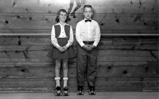 A black and white photo of a young boy and girl in a roller skating rink dressed up and wearing black roller skates.