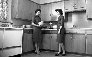 A black and white photo of two women in 1950s style dresses standing in a kitchen. 