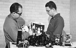 A black and white photo of two men with a collection of cameras and photos. 