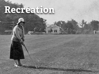 A black and white image of a woman in a long skirt, sleeved shirt, and hat playing golf. 