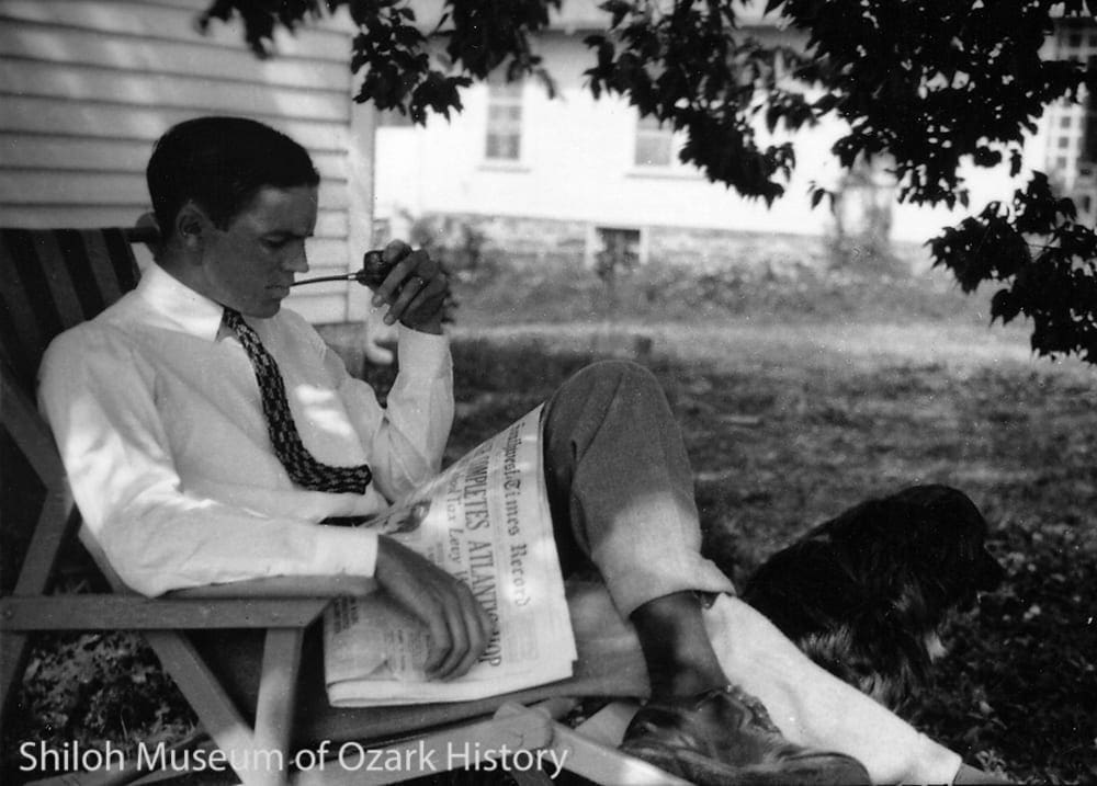 A black and white image of a man sitting outside in a chair reading the news.