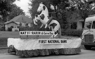 A black and white photo of a parade float with a fake rearing horse and a woman sitting on the horse. 