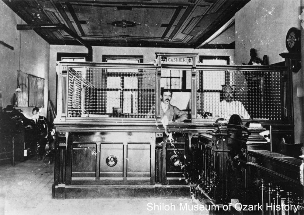 A wooden bank counter in 1895 with an iron grill and two bankers behind the counter.