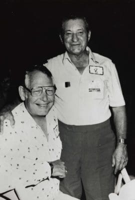 Black and white photo of two men, one is standing and one is seated. They are smiling.
