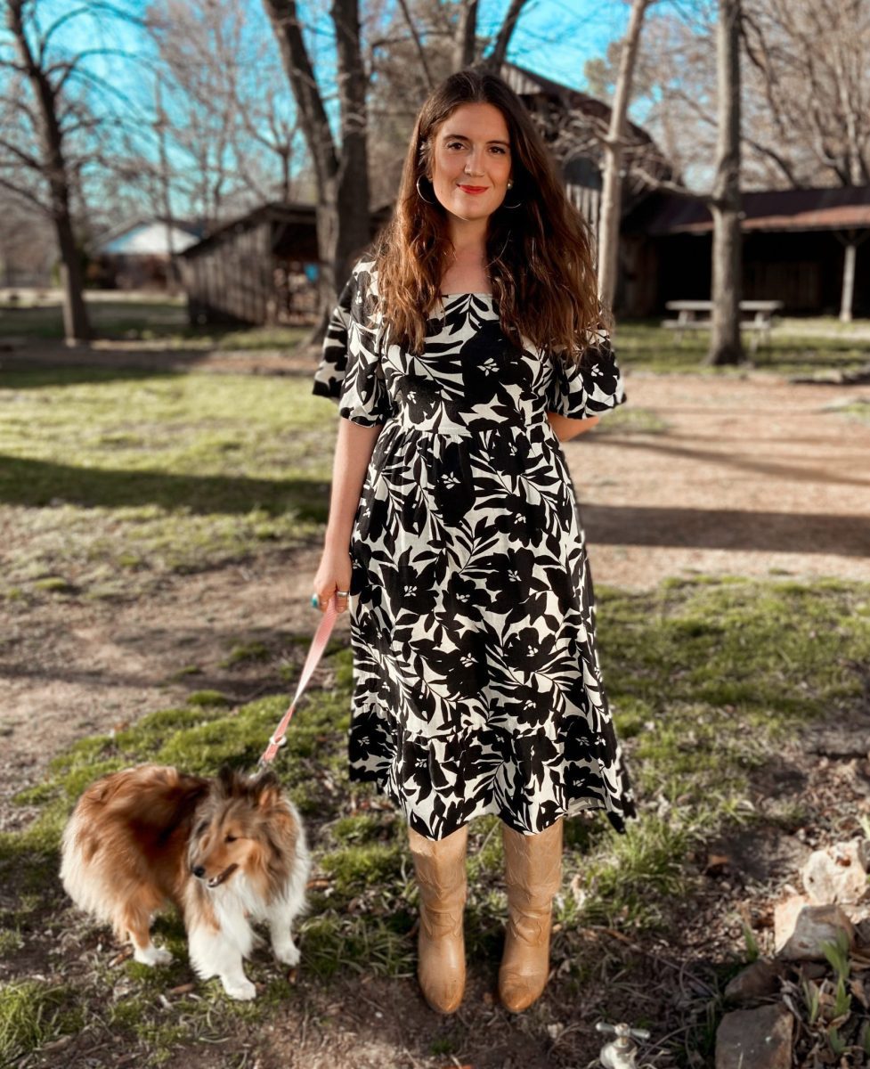 Woman standing in front a barn with long dark hair, black and white dress, holding the leash of a dog.