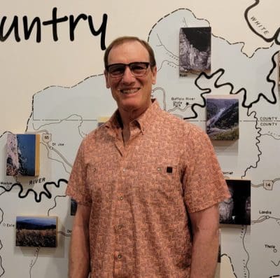 Man wearing glasses and patterned orange shirt standing in front of wall with a map of the Buffalo river and photographs on top of the map.