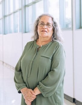 Woman with green blouse, glasses, long gray hair, standing in front of windows.