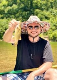 Man in sunglasses with a river behind him. Holding up a fish for the camera.