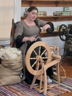 Woman in dress sitting behind spinning wheel next to a bag of cotton.