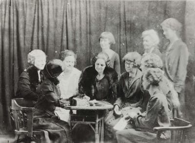 Black-and-white photograph of 10 women, all wearing dresses, two with fur collars and one is wearing a hats. Six are sitting around a small table looking down at it with the woman seated on the left appearing to write on something on the table while another woman is either pointing or writing as well. Three women, two of whom are blurred, stand to the right. A dark curtain hangs in the background.