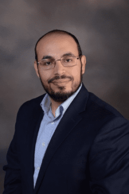 Photo of a man with a dark beard, dark, short-cropped hair and glasses in a navy blazer and light blue collared shirt. 