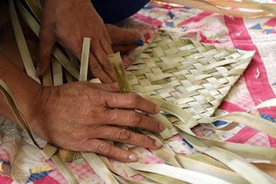 Close up image of hands weaving a mat with flat, organic fibers.