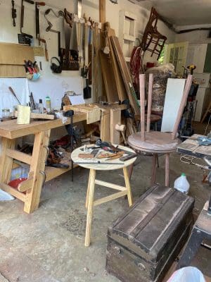 Image of woodworking shop consisting of a work bench, and three round-top tables in different states of construction, a trunk in the foreground and tools, boards, and a hair all hanging from a wall and an open door in the back of the room.