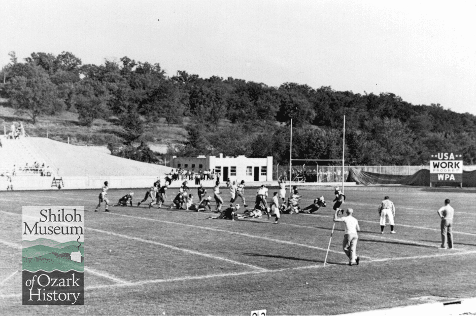 Football players on a field with a tree-covered hill, bleachers, building and a sign that reads, 
