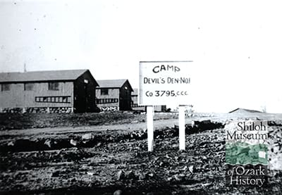 Black-and-white image of a rocky, dirty field with a dirt road and three camp buildings with ladders hanging horizontally and windows on each side. In the foreground is a white, wooden sign that reads, “Camp Devil’s Den-No. 1, Co. 3795, C.C.C.” At the bottom right is the Shiloh Museum of Ozark History logo.