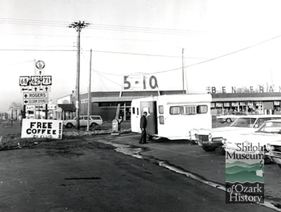Black-and-white image of a highway intersection with road signs and a painted handmade sign that reads “Free Coffee, CB Club.” To the right of the road is a parking lot with cars and a camper trailer with a man entering. Behind the trailer is a building with an arch façade with a sign above the roof that reads, “5-10.” To the right on the building’s roof are the letters “Ben Fran” discernible. Road signs point to directions to Rogers and Siloam Springs and to highways 68, 62, and 71.