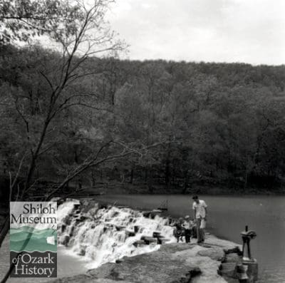 Black and white photograph of a distant shot of young girl and boy walking with man on flat capstone at end of curved, multi-tiered rock dam with water flowing over with sparsely leafed trees in the background.