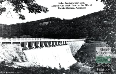 Black-and-white image of a dam with light-colored bricks on each side and water flowing through columns in the center. Tree covered hills are behind it. A Shiloh Museum of Ozark History logo is in the bottom right corner.