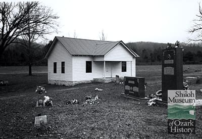 Black-and-white image of a small, one-story light-color clapboard church on stone foundation with portico supported by two columns over porch floor. Grass and tombstones in foreground, and grass, trees, and tree-covered hills are in the background.
