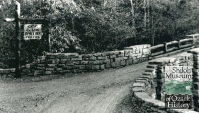 Black-and-white image of a stone bridge with log railing with a dirt road leading up to it and trees in the background. At the entrance of the bridge is a sign made of log poles that reads, “C.C.C. Co. 3195, Devil’s Den, PROJ. SPS.” Shiloh Museum of Ozark History logo is at the bottom right.