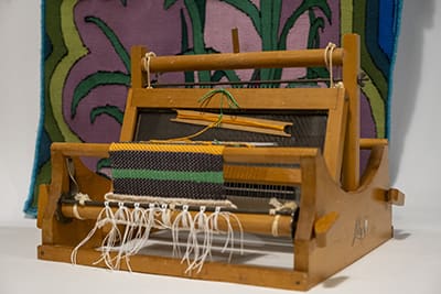 A wooden loom with an orange, brown, and green-striped scarf with a white trim and tassels hanging from it as it’s being woven. Behind it is a tapestry of various shades of green and mauve with a design involving stalks and leaves.