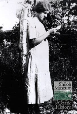Black-and-white photo taken outdoors of a young woman in light-color dress standing in tall grass, reading letter with a fence, brush, and foliage in background. A Shiloh Museum of Ozark History logo is on the bottom right of the photo.