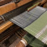 Detail image of a large, wood-frame loom with partially woven green, pink, and white fabric.