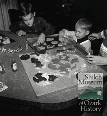Black-and-white photo of two elementary school-aged boys and two adult hands visible at a table with cookie sheets, cookie cutters and shaped dough.