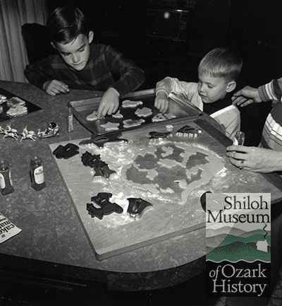 Black-and-white photo of two elementary school-aged boys and two adult hands visible at a table with cookie sheets, cookie cutters and shaped dough.