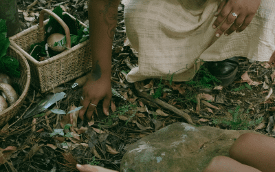 Closed up of the edge of dress, shoe and hands reaching toward something n the ground next to baskets with mushrooms.