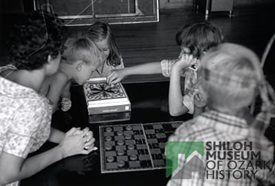 Black-and-white photo of two boys, two girls, and a young woman sitting at a table playing games with a checkerboard in the foreground.