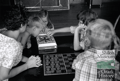 Black-and-white photo of two boys, two girls, and a young woman sitting at a table playing games with a checkerboard in the foreground.