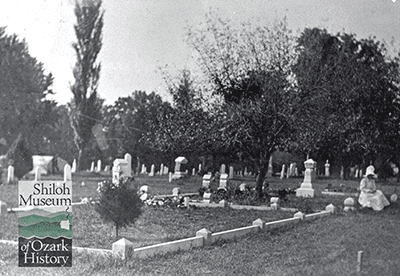 Old black-and-white photo of a cemetery with plots bordered by a low, concrete fence and trees in the background and a woman sitting on the low fence wearing a long white dress and bonnet, her face not visible.