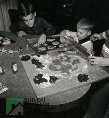 Black-and-white photo of two elementary school-aged boys and two adult hands visible at a table with cookie sheets, cookie cutters and shaped dough.