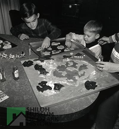 Black-and-white photo of two elementary school-aged boys and two adult hands visible at a table with cookie sheets, cookie cutters and shaped dough.