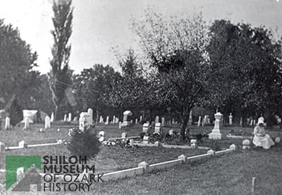 Old black-and-white photo of a cemetery with plots bordered by a low, concrete fence and trees in the background and a woman sitting on the low fence wearing a long white dress and bonnet, her face not visible.