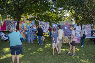 Photo of people milling about on a lawn beneath trees and a stretched clothesline of quilts hanging from it.