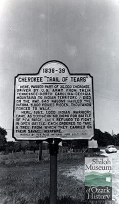 Black-and-white photo of a metal marker with words on the side of a gravel road with a car in the distance.