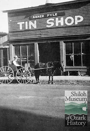 Man in carriage being pulled by horse in front of a building with a flat roof, two large windows and a sign overhead that reads, “Abner Pyle Tin Shop.”