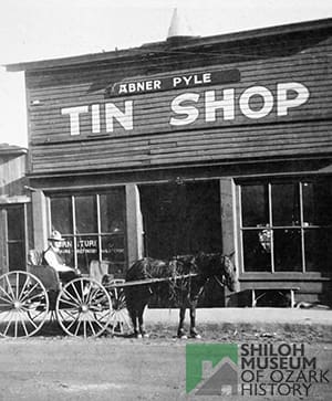 Man in carriage being pulled by horse in front of a building with a flat roof, two large windows and a sign overhead that reads, “Abner Pyle Tin Shop.”