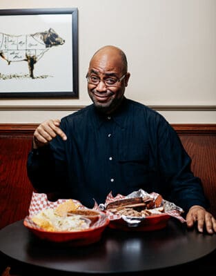 A man wearing a black shirt sitting at a table while smiling and pointing at two plate baskets of barbecue.