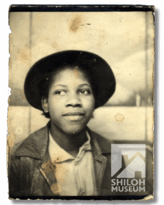 A young person in a worn, vintage photo, wearing a hat and leather jacket, looks calmly to the side. The image is faded with visible stains.