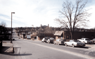 A quiet, suburban street with vintage cars parked along a row of small businesses. Leafless trees line the road under an overcast sky, creating a nostalgic tone.