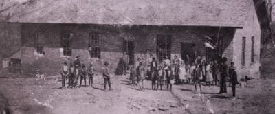 Black and white image of Henderson School circa 1875. Numerous Black children seen from a distance in front of the school.