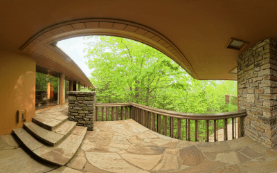 Curved stone patio with wooden railing overlooks lush green trees. Rustic stonework and warm tones create a serene, natural retreat vibe.