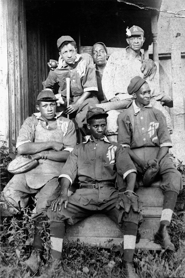 Historic black and white photo of a vintage baseball team. Six young men in uniforms and caps sit on steps, holding bats and gloves, conveying camaraderie.