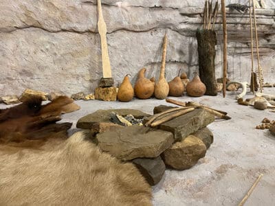 Recreation of a primitive camp: stone fire pit, animal skins, dried gourds, wooden tools, and spears against a textured cave-like wall. Rustic and historical.