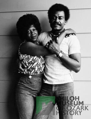 A man and woman stand close together, embracing in front of a textured wall. The woman wears a patterned top and jeans, the man a light shirt and jeans. Both look directly at the camera, conveying a sense of warmth and intimacy. Text reads: