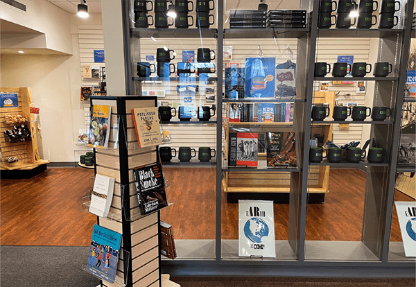 Books and souvenirs on display in a store with wooden flooring and bright lighting. Shelves feature mugs, posters, and varied book titles.