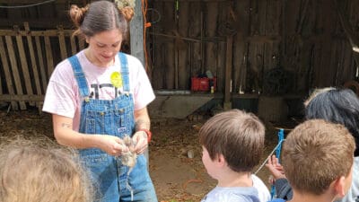 A person in overalls and a pink shirt interacts with a group of children, holding a small object. The setting appears rustic and educational, with a warm tone.